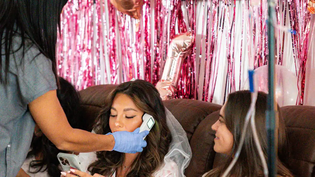 A nurse checks a guest's temperature before her drip while a friend looks on from the couch