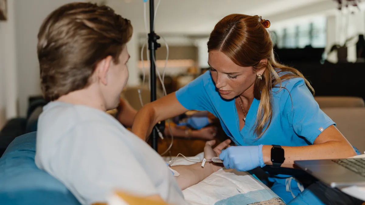 A Drip Lab nurse in blue scrubs starts an IV for a client relaxing at home