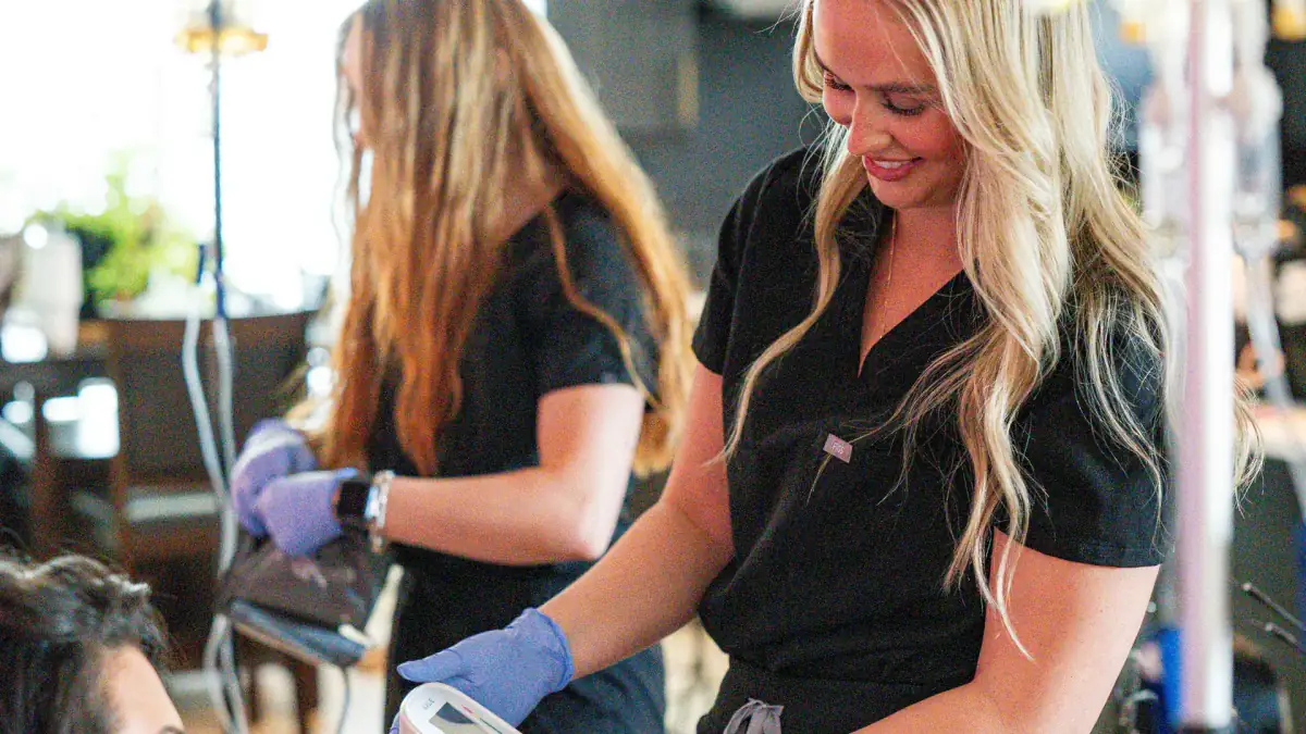 Two Drip Lab nurses in black scrubs check on a client during a home visit