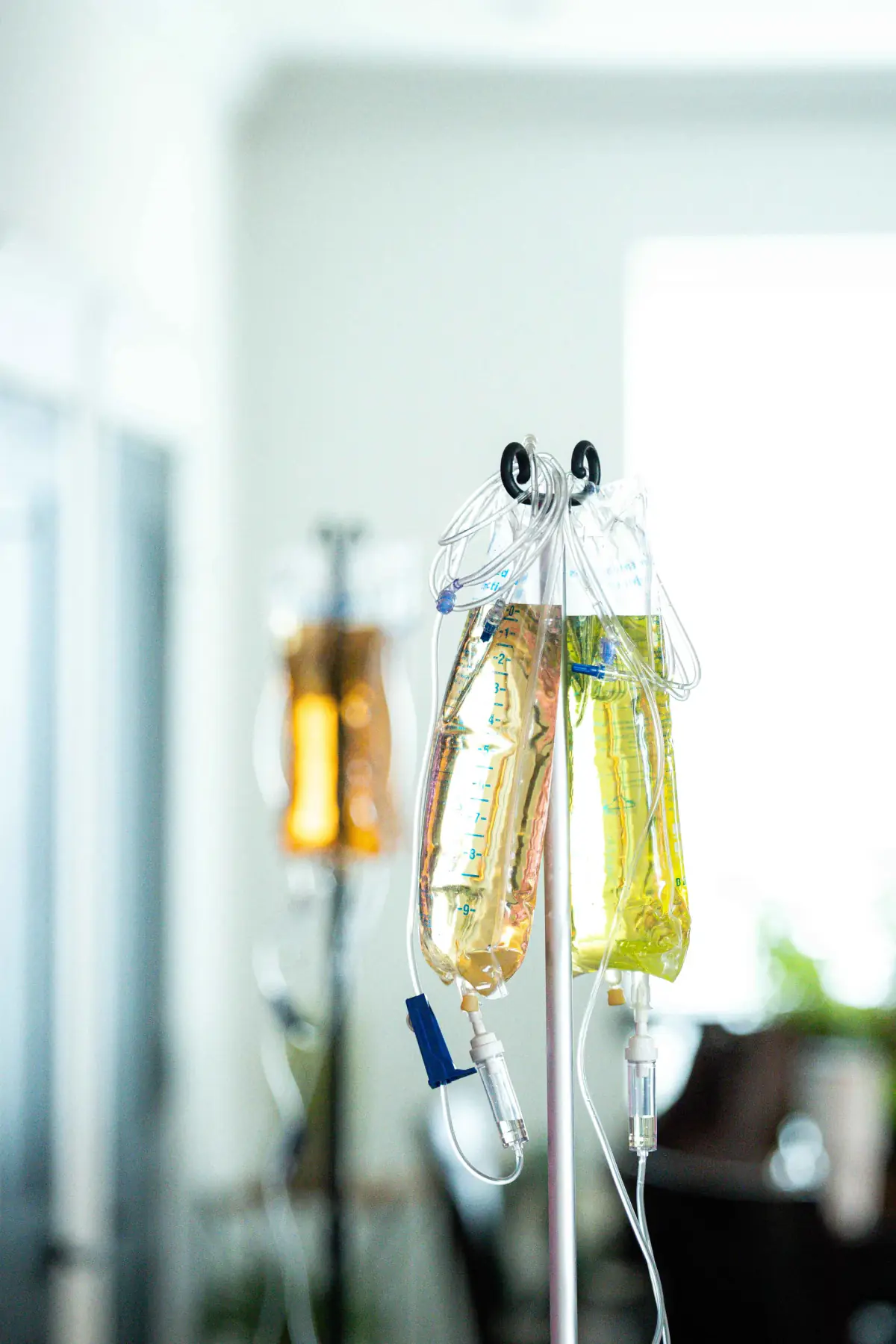 Two IV bags, one amber and one yellow, hang together from an IV pole in a sunny room